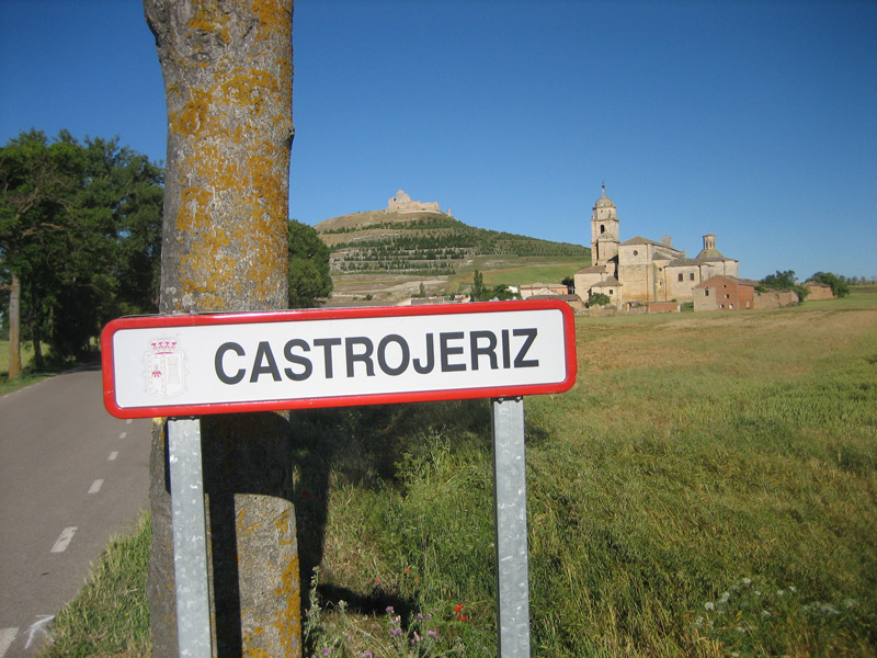Ortsschild „Castrojeriz“ am Weg, dahinter die Kirche und die Burgruine auf dem Hügel