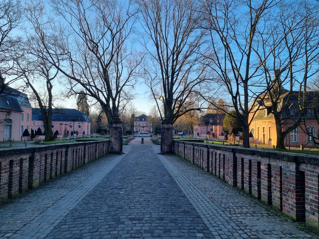 Gepflasterte Brücke zwischen Backsteinmauern, kahle Alleebäume, im Hintergrund Schloss Wickrath