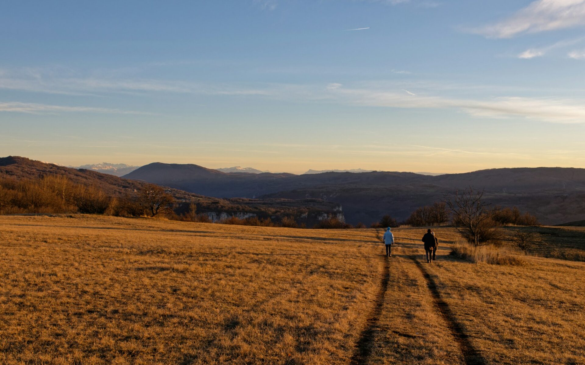 Zwei Wanderer auf einer Wiese mit Fahrspur, dahinter Hügelketten im Abendlicht